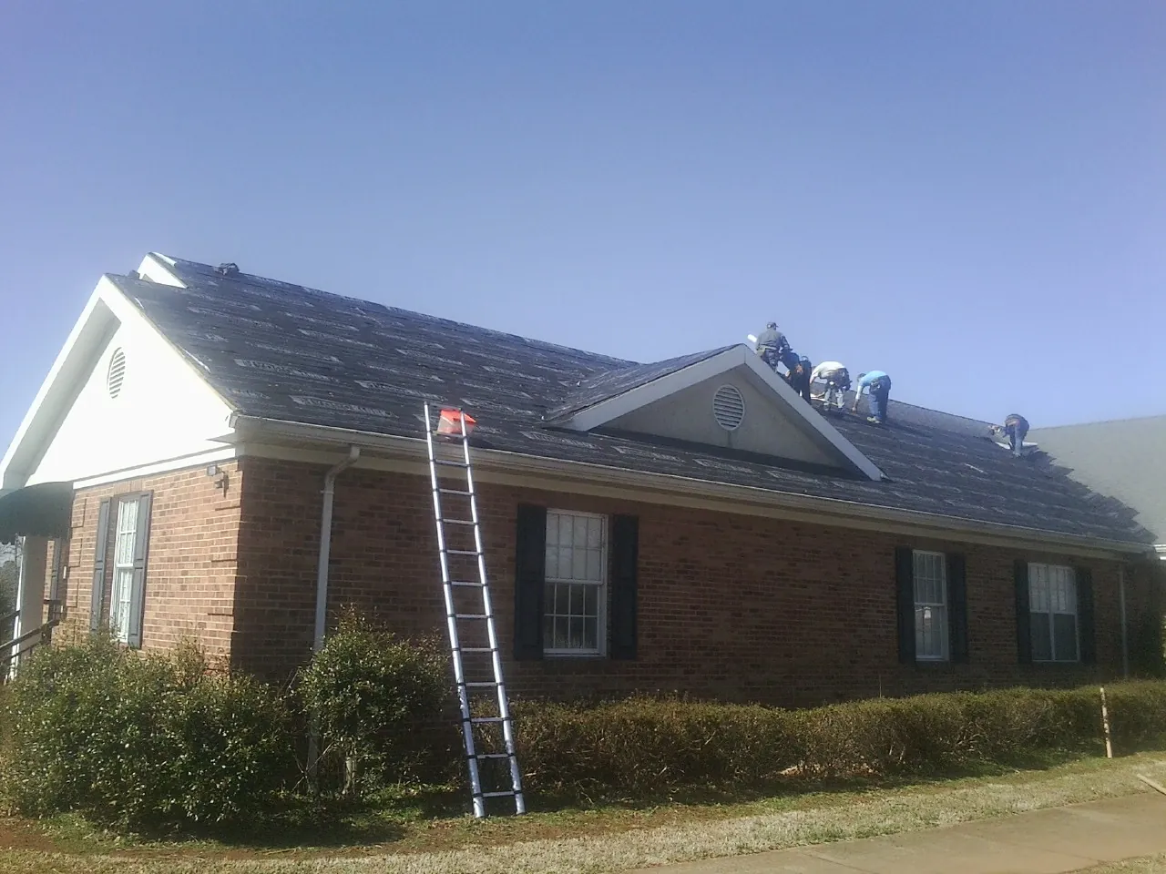 Crew tearing shingles off a brick ranch roof with a ladder set — Metal Roof Repair work by our crew in Castle Hayne