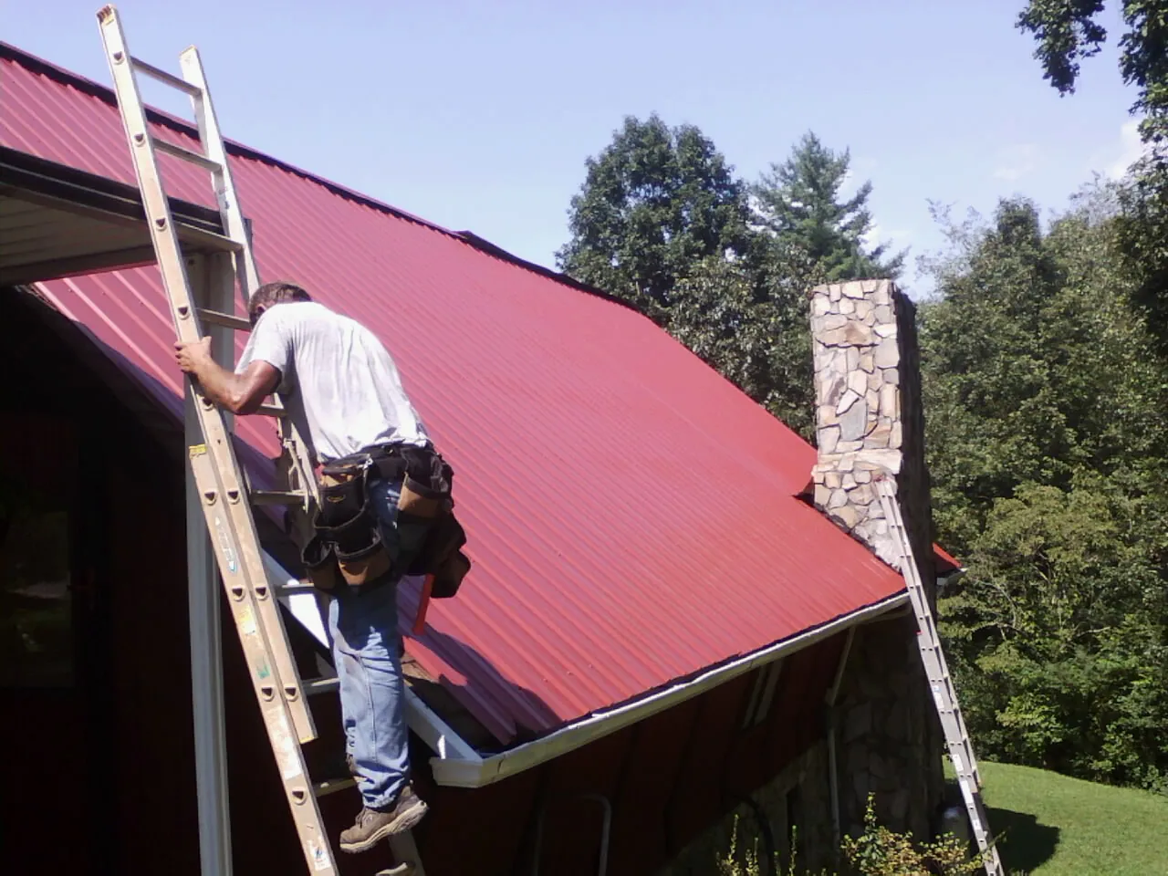 Roofer on a ladder beside a red metal roof and stone chimney — Soffit & Fascia Repair by Roofing Brothers in Castle Hayne